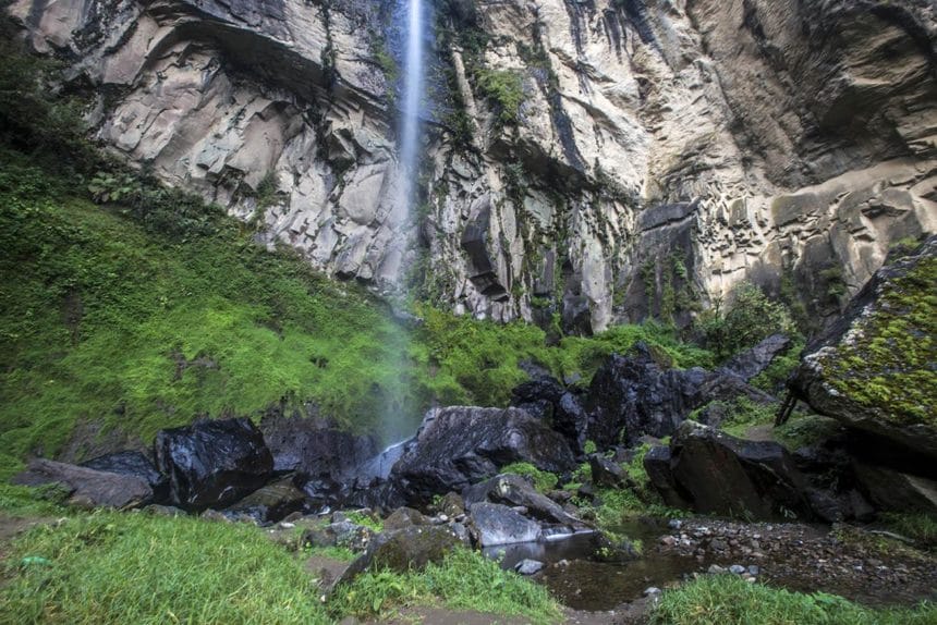 A tall, narrow waterfall cascades down a rocky cliff, surrounded by lush green moss and stones, creating a serene, natural scene.