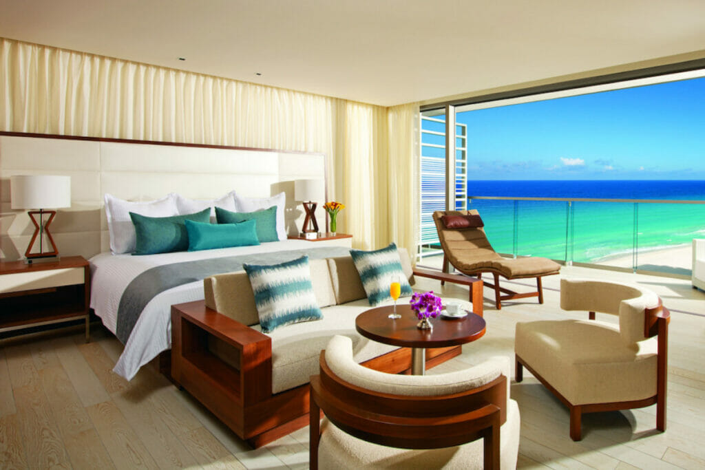 A large room with a bed surrounded by chairs and a table with a view of the beach in Secrets the Vine