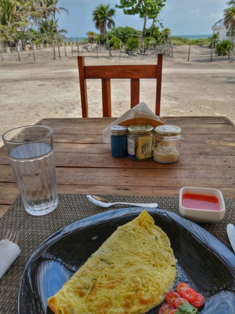 A beachside table features an omelet, fresh juice, and condiments, offering a serene view of palm trees and the distant ocean.
