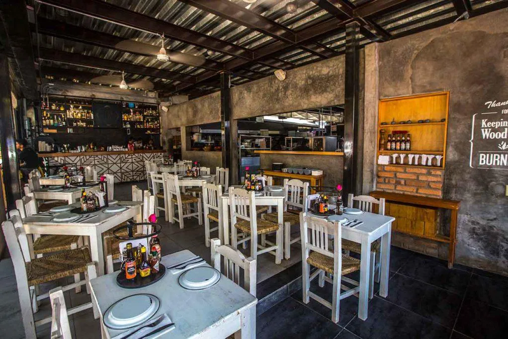 Chic, rustic interior of La Rustica restaurant featuring white wooden tables, woven chairs, exposed brick, and a backlit bar fully stocked with spirits.
