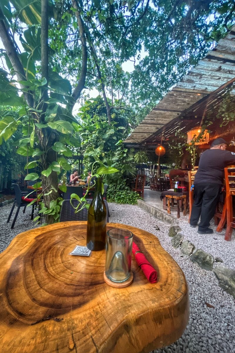 A rustic outdoor restaurant with lush greenery surrounds wooden tables. A person stands near a sheltered dining area, creating a cozy, natural atmosphere.