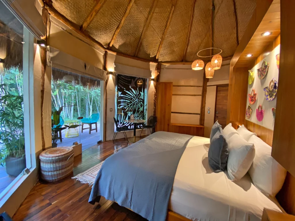 A cozy, rustic bedroom features a thatched ceiling, colorful wall art, wooden decor, and a view of greenery through large glass doors.