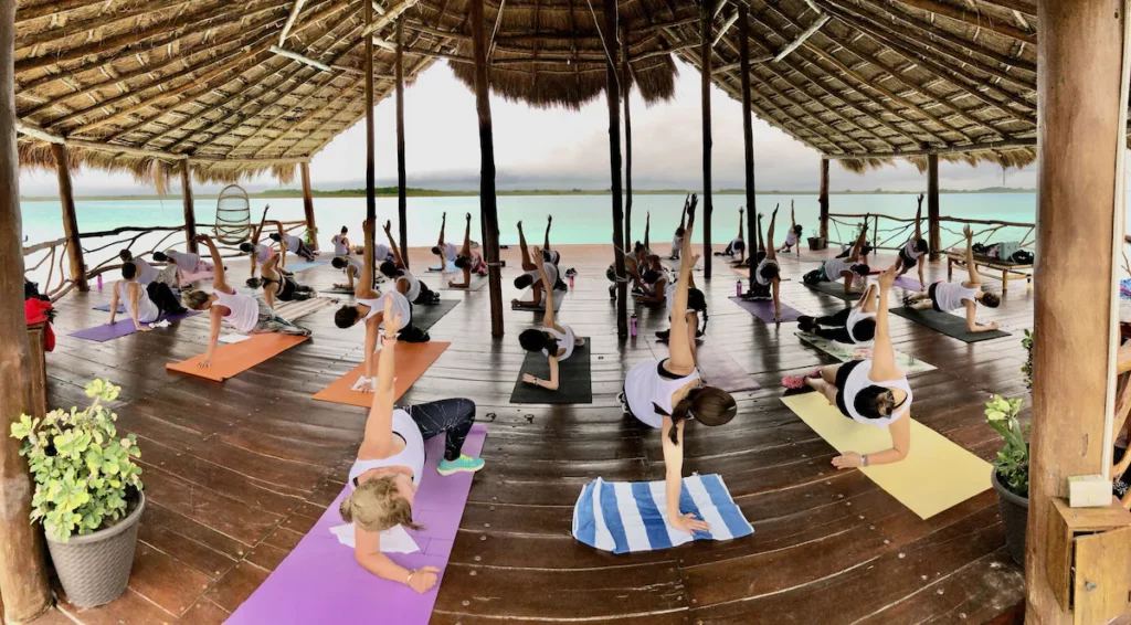 A group of people practice yoga under a thatched roof pavilion near a turquoise lake, surrounded by lush greenery and wooden railings.