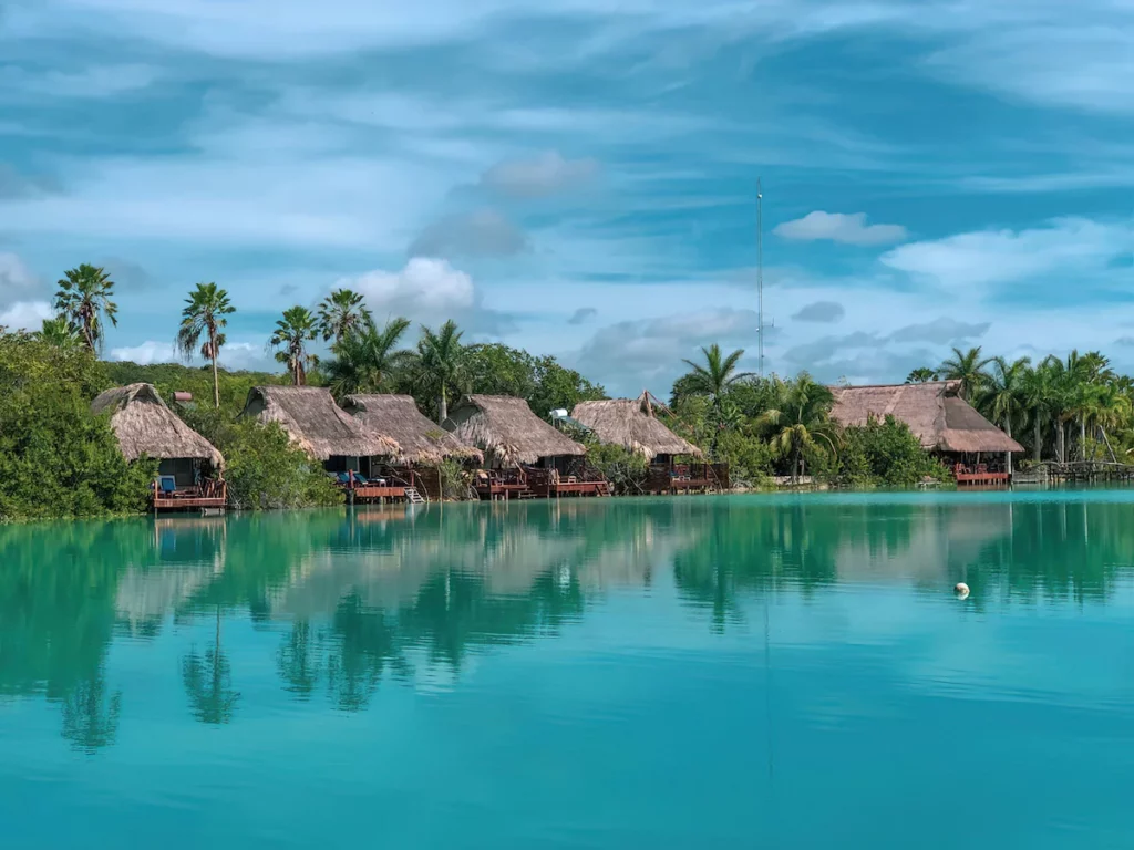 Thatched-roof huts lined along a clear turquoise lake, surrounded by lush palm trees under a vibrant blue sky. Tranquil, scenic tropical setting.