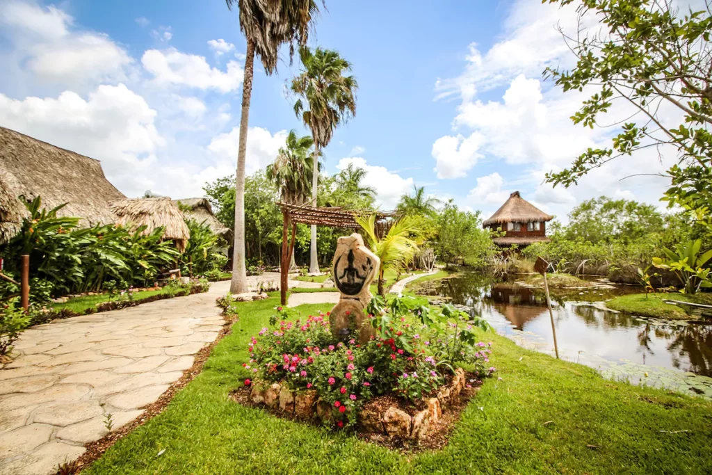 Lush garden with stone pathways, vibrant flowers, palm trees, and a thatched-roof structure beside a tranquil pond under a bright blue sky.