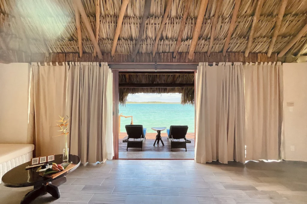 Tropical hut interior with thatched roof, overlooking a deck with two loungers facing a turquoise sea, creating a tranquil, seaside retreat ambiance.