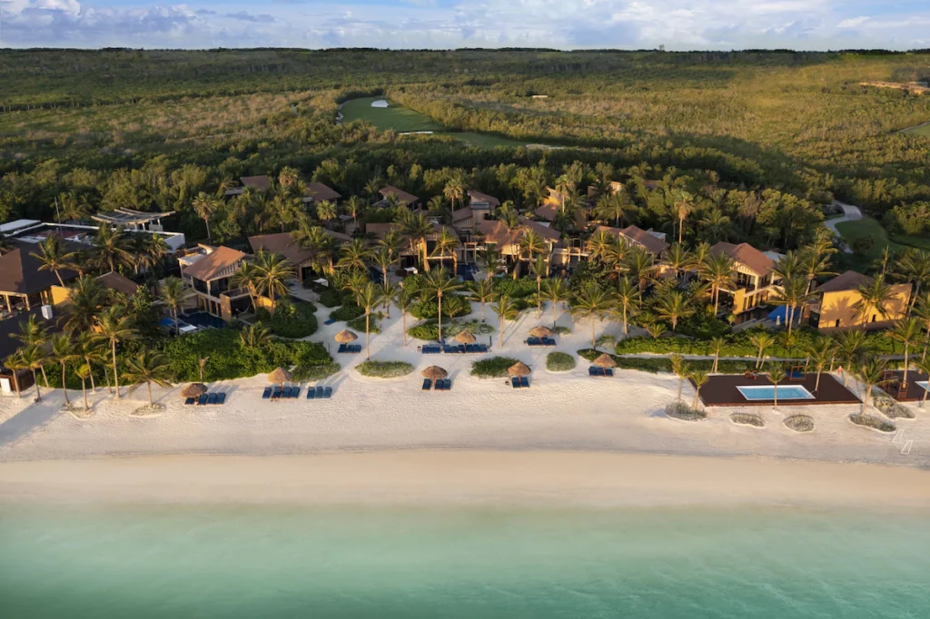 Tropical resort with sandy beach, palm trees, lounge chairs, and thatched umbrellas. Lush green forest surrounds the buildings. Calm waters line the shoreline.