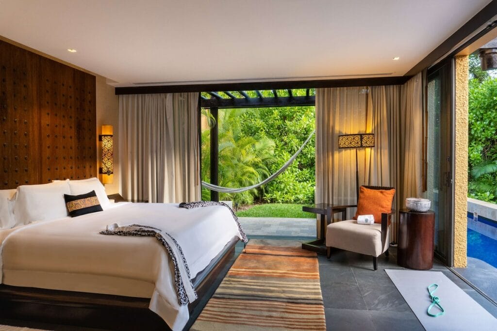 A luxurious bedroom opens to a tropical garden with a hammock and pool, featuring modern decor and large windows for natural light.