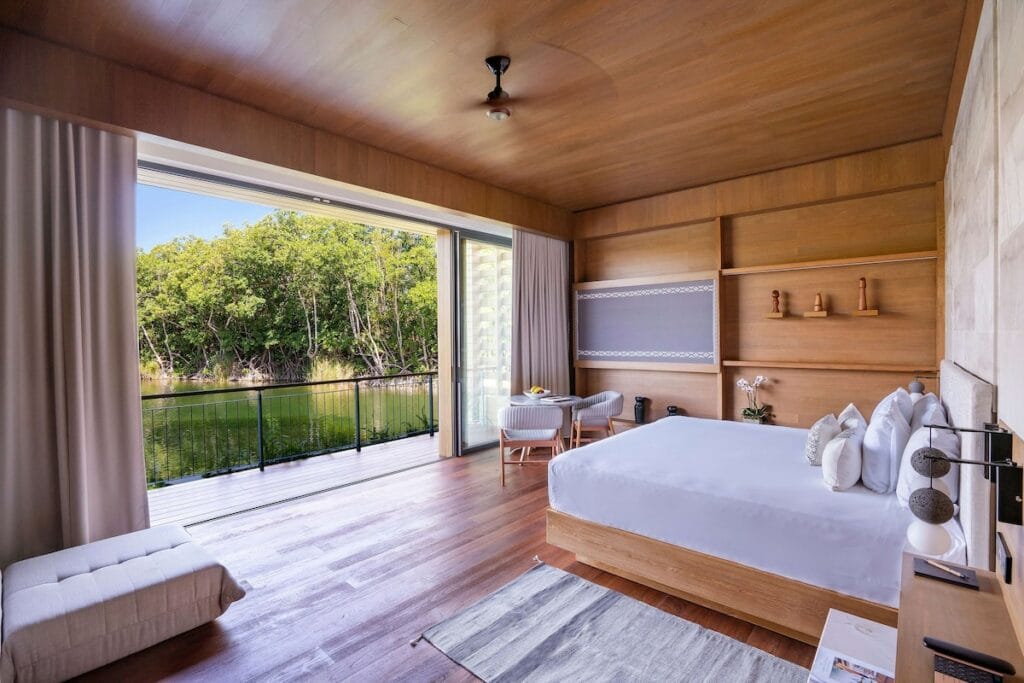 A serene, modern bedroom with wooden accents, large bed, and an open view of a lush forest and tranquil water outside.