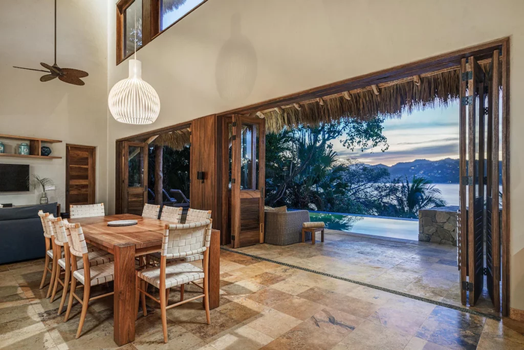 A rustic indoor dining area with woven-back chairs and a wooden table opens up to a thatched patio with a panoramic ocean view and infinity pool at sunset.