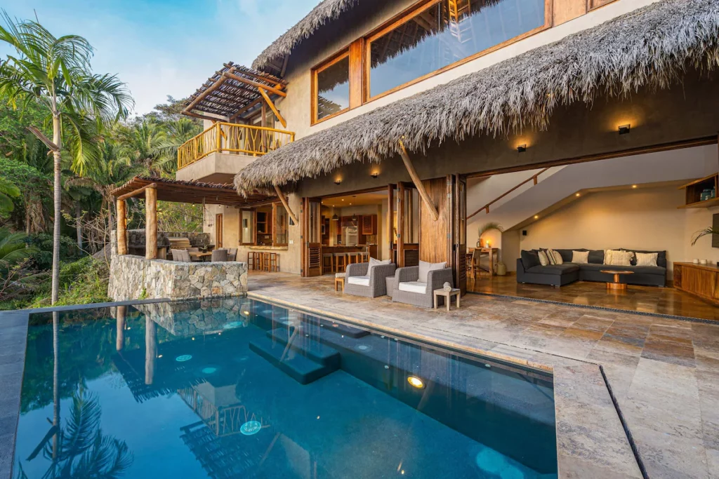 The exterior of a tropical villa features an open living space leading to a stone-tiled patio and serene infinity pool surrounded by lush palm trees.
