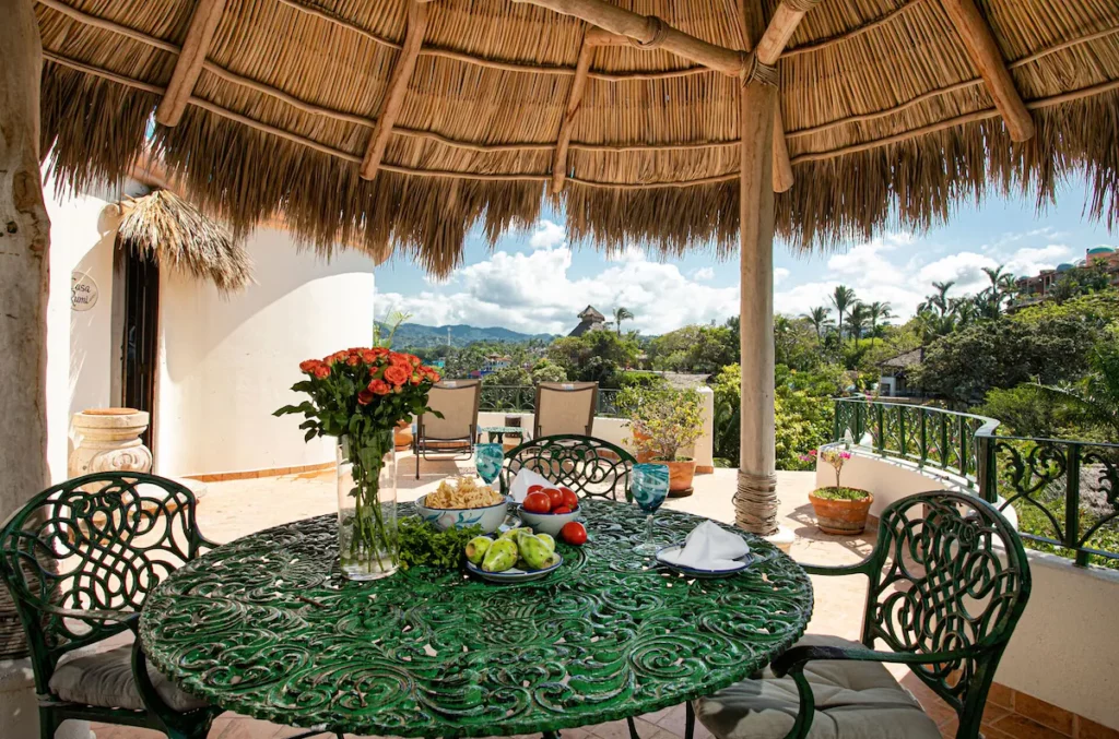 Outdoor terrace under a thatched palapa with a green wrought-iron dining table, surrounded by tropical plants and views of nearby hills and rooftops.