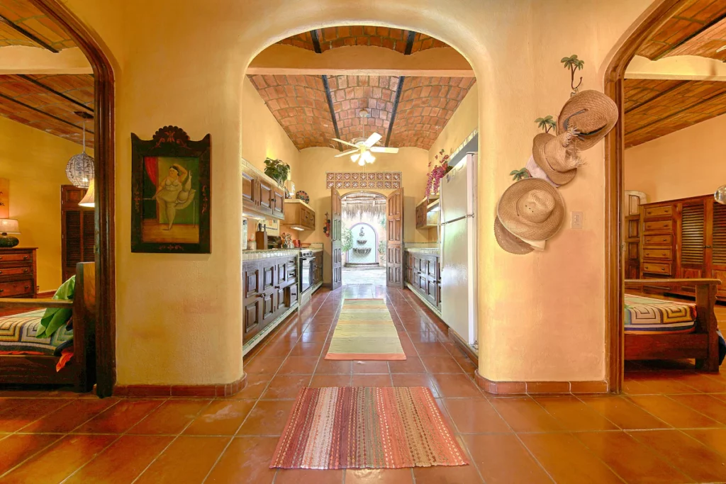A warm and rustic hallway with archways, clay tile floors, and a vaulted brick ceiling leads to a wooden front door and courtyard fountain. Straw hats hang on the wall, and the space feels homey and traditionally Mexican.