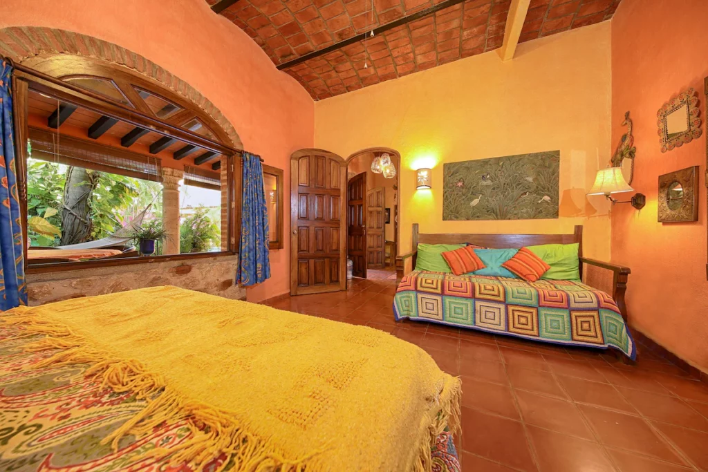 A cozy bedroom with warm orange walls, colorful patterned bedding, and arched windows overlooking lush greenery and a hammock outside. The room features hand-painted artwork, wooden furniture, and charming rustic decor.