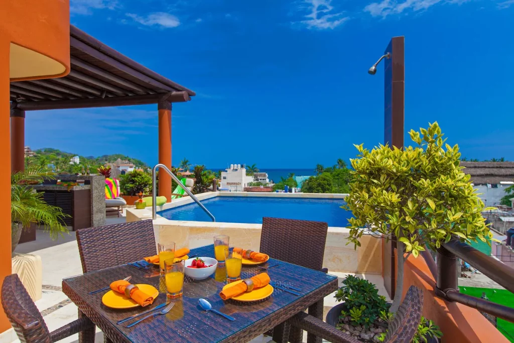 Rooftop breakfast setup beside an infinity pool, with a wicker dining table set for four and panoramic views of the ocean and vibrant blue sky. Tropical plants and colorful textiles bring in vacation vib