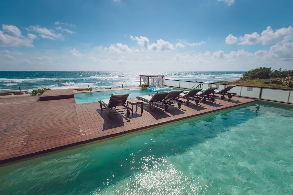 Ocean view features an infinity pool, wooden deck with loungers, sunbeds, and a bright blue sky with scattered clouds.