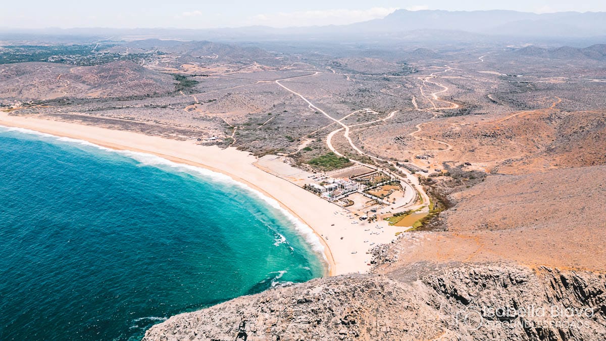 Aerial view of a coastal desert landscape with a pristine beach, turquoise ocean, and a small resort. Rocky terrain surrounds the area.
